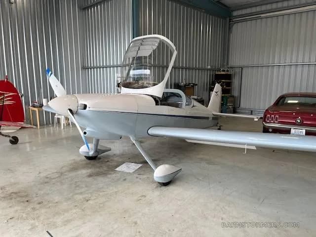 A light blue and white single-engine aircraft with its canopy open, parked inside a hangar next to a red classic car.