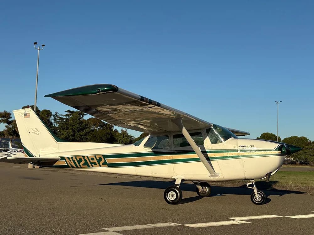 A white Cessna 172 with green and gold stripes parked on an airfield with trees in the background.
