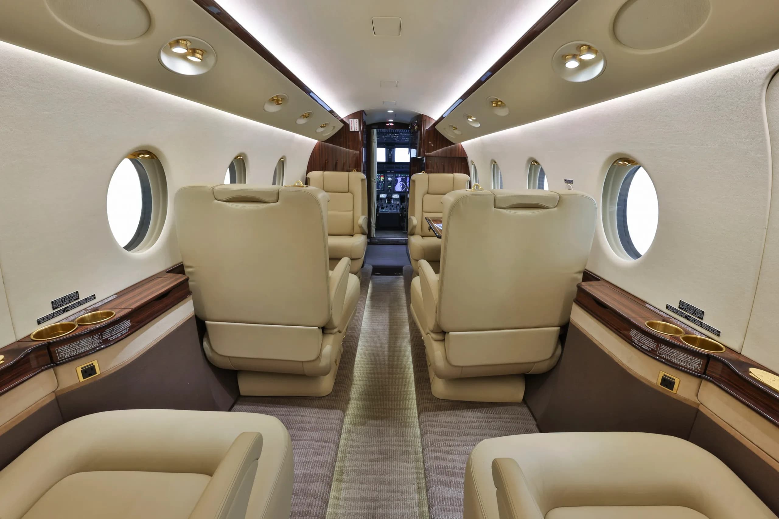 A view down the aisle of a private jet cabin with beige leather seats and polished wood accents.