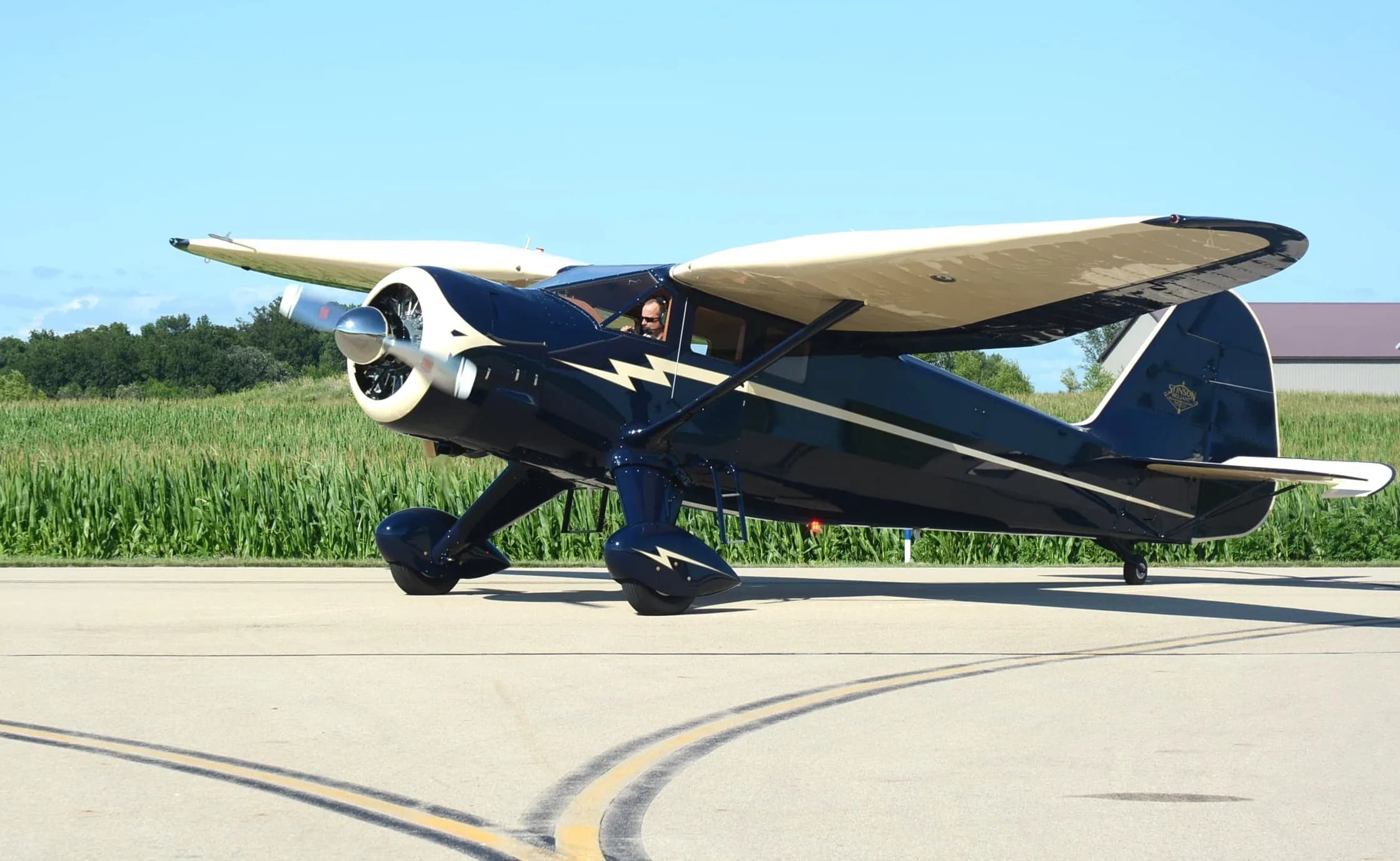 A dark blue and cream vintage aircraft with a lightning bolt design is taxiing on a tarmac next to a green field.