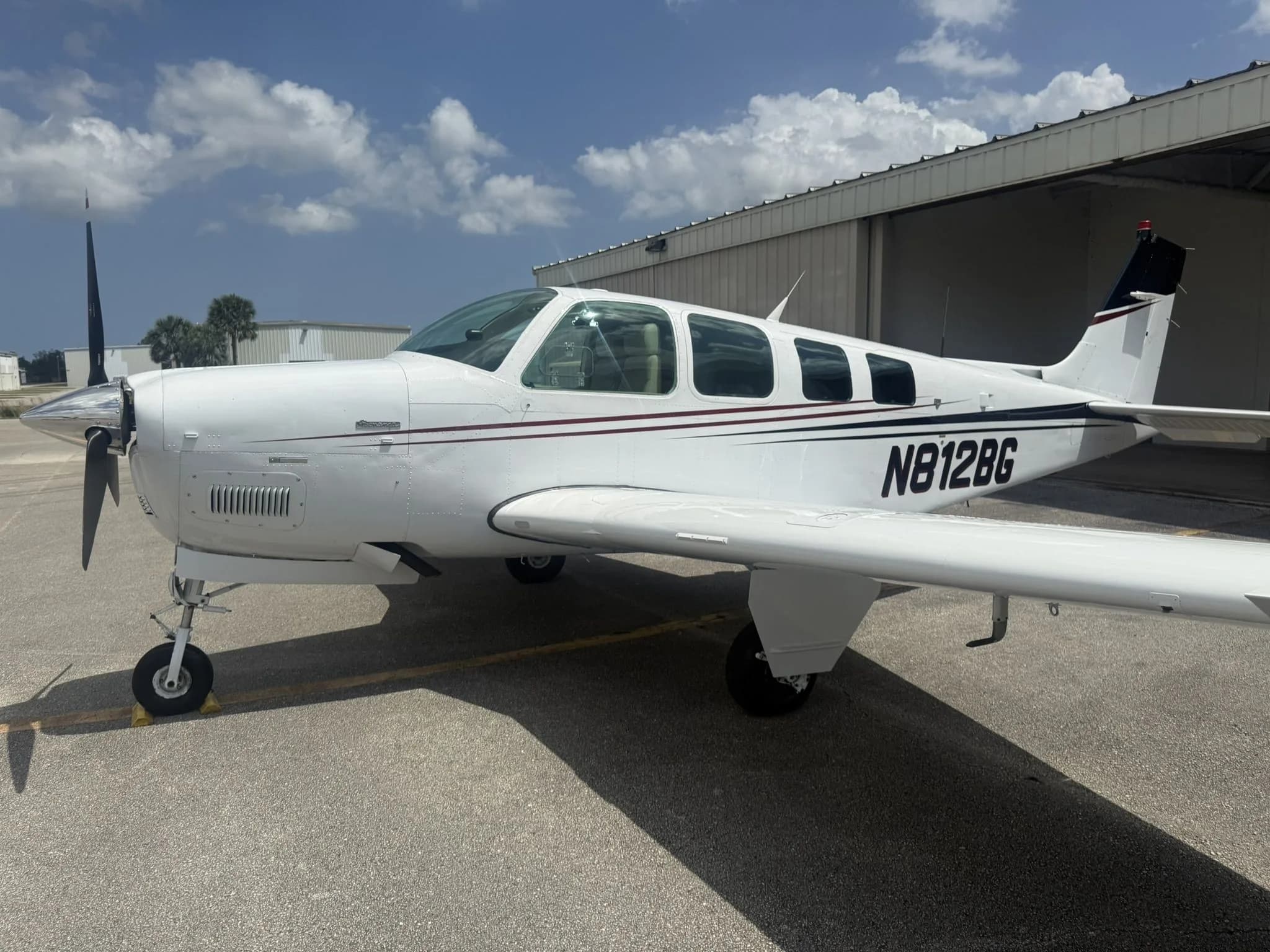 White Beechcraft Bonanza N812BG with dark red and black stripes parked on an airfield tarmac.