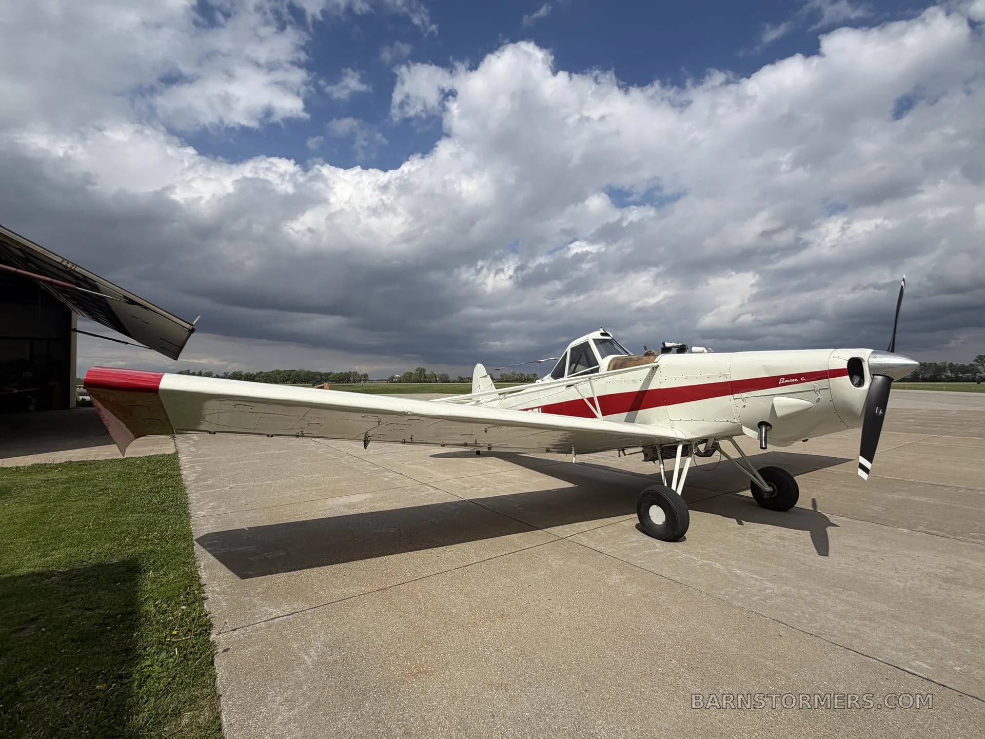 A white and red agricultural aircraft, seen from the front-right side, parked on an airfield under a partly cloudy sky.