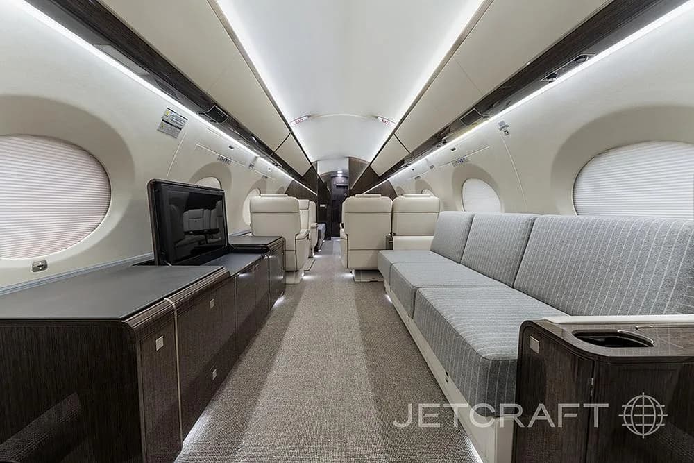 A private jet cabin with a grey fabric sofa on the right and a dark wood cabinet with a pop-up television on the left.