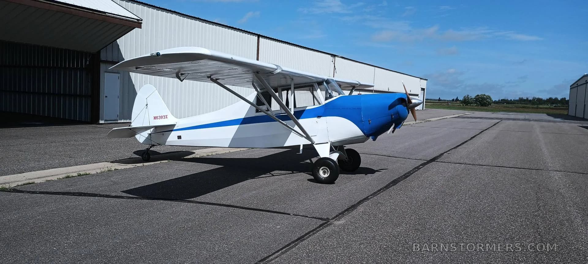 A white and blue single-engine taildragger aircraft, N6393X, parked on an asphalt ramp in front of hangars.