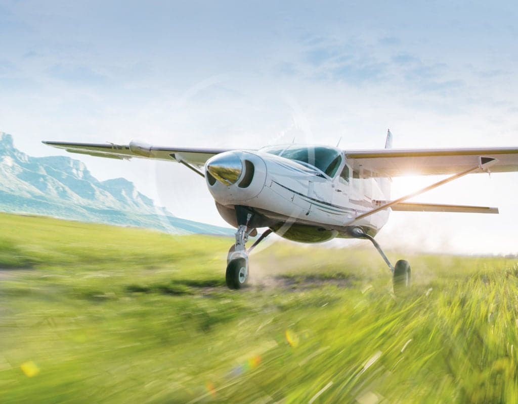 A Cessna Caravan taking off from a grassy field with mountains in the background.