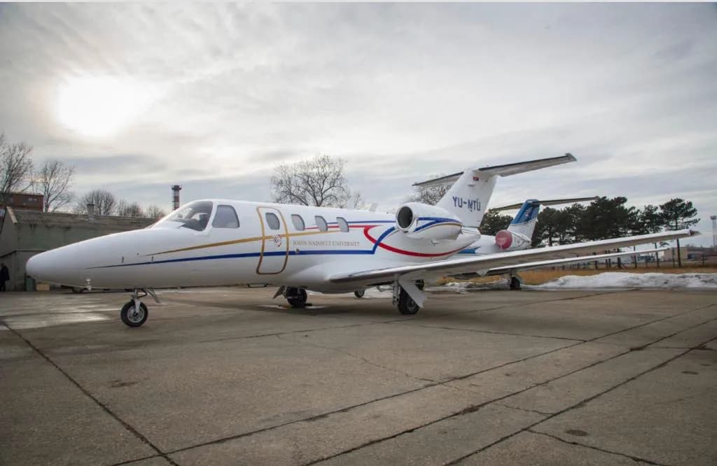 A white business jet with blue, red, and gold stripes, and "JOHN NAISBITT UNIVERSITY" text, parked on an asphalt apron under a cloudy sky.
