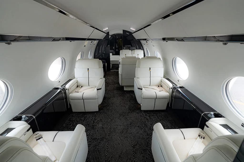 The luxurious cabin interior of a business jet featuring white leather seats and dark patterned carpet.