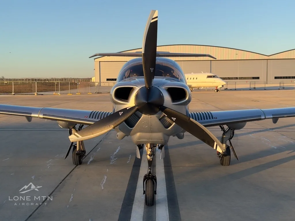 A head-on view of a silver Diamond DA50 aircraft with its propeller blades visible, parked on an airfield.