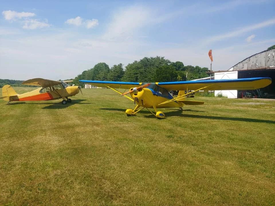 Two light aircraft, one yellow and orange and one yellow and blue, parked on a grassy airfield next to a hangar.