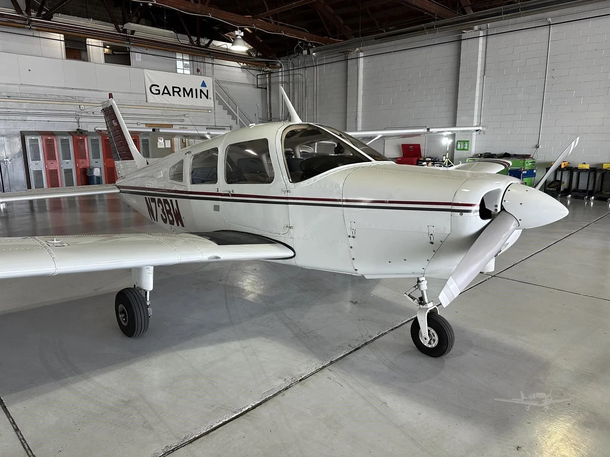 A close-up front-right view of a white single-engine aircraft with maroon and black stripes, registration N73BW, in a hangar.