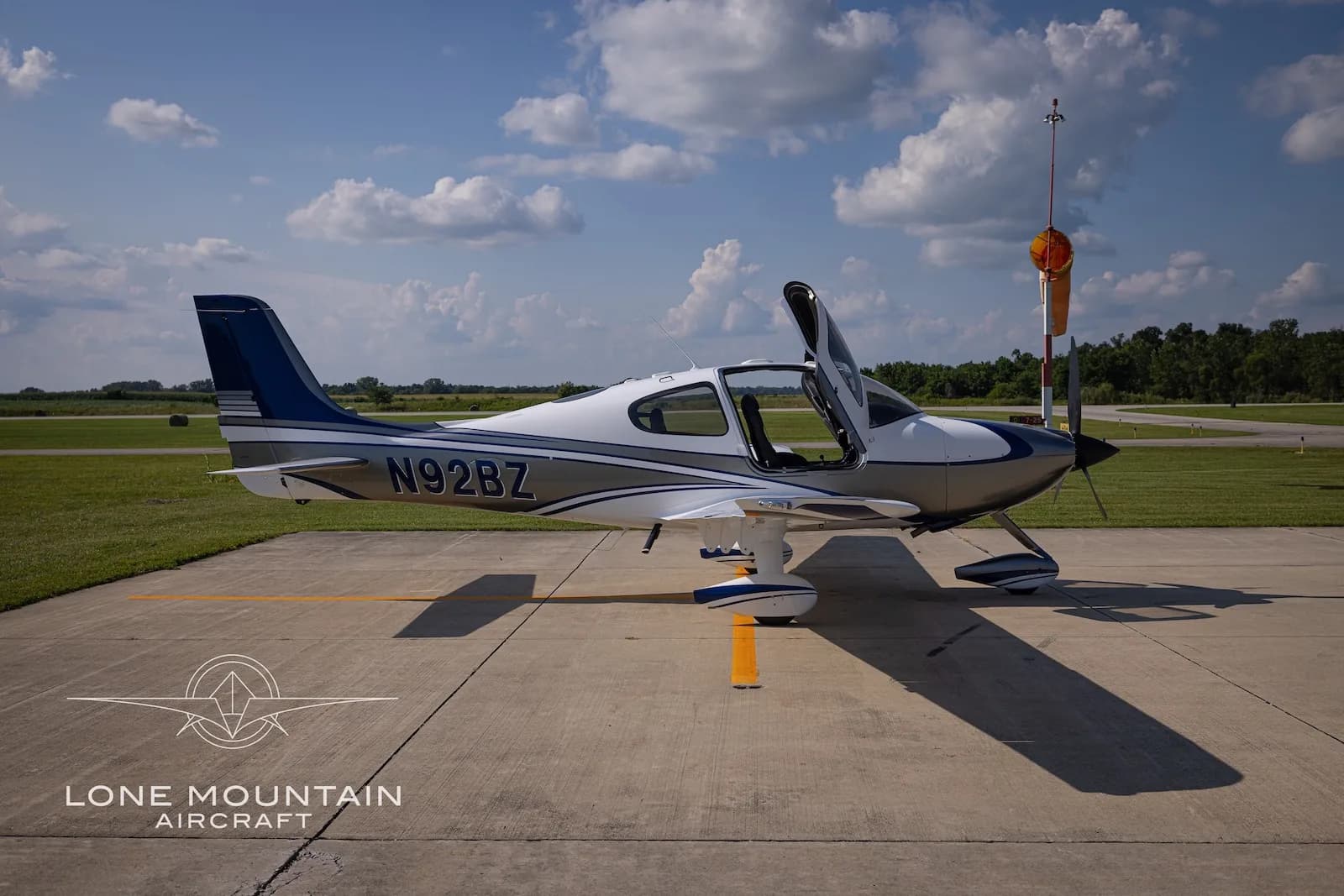 A side view of the white and silver Cirrus SR22 aircraft N92BZ with blue accents, parked on an airfield with its doors open.