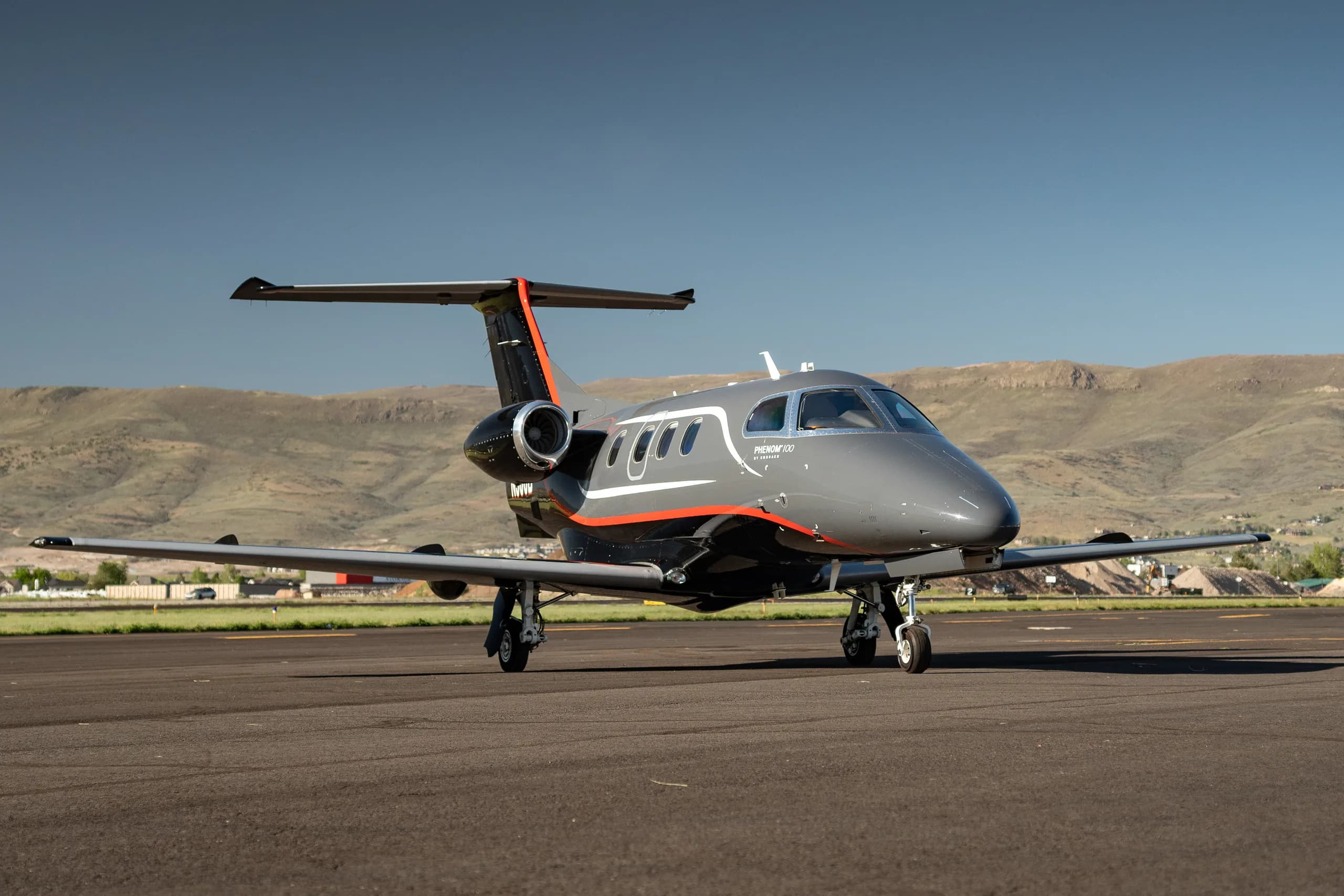 A dark gray and black Embraer Phenom 100 with red and white stripes, registration N506B, parked on a runway, viewed from the front.