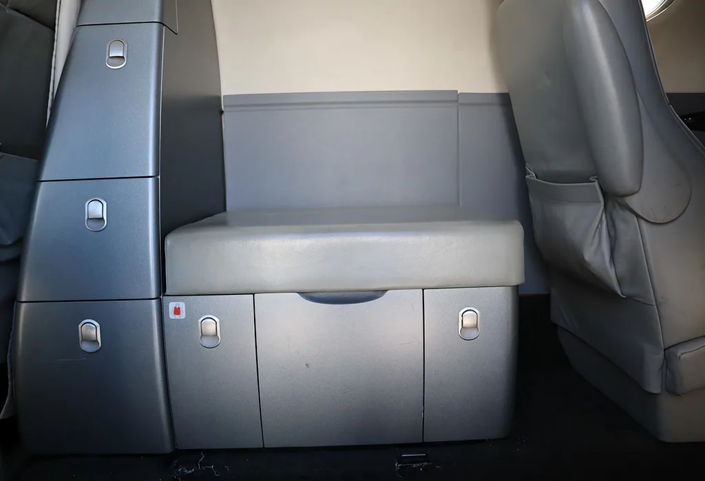 A compact gray storage unit or lavatory area with a padded top and drawers next to a seat in the aircraft cabin.