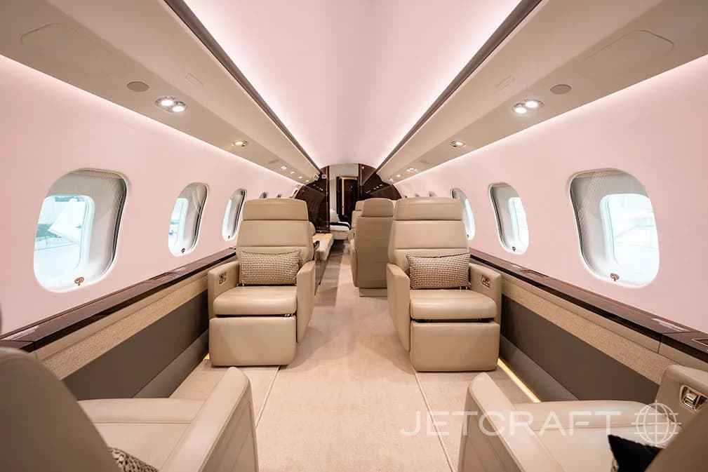 The luxurious cabin interior of a business jet, showing two rows of beige leather seats facing each other, with ambient lighting.