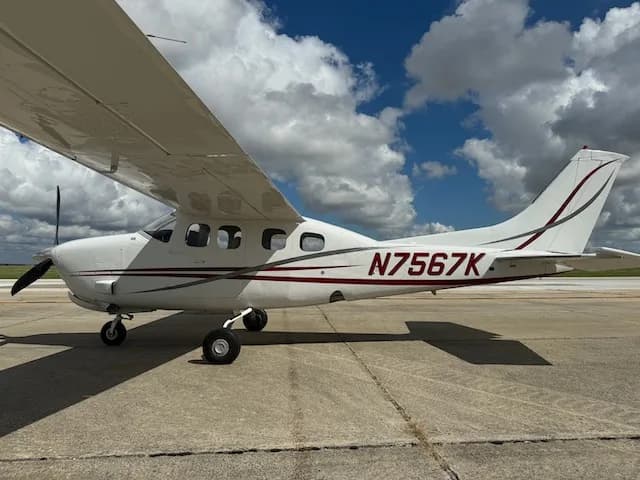 A side view of a white Cessna 206 with red and grey stripes, clearly showing the tail number N7567K, parked on an airfield.