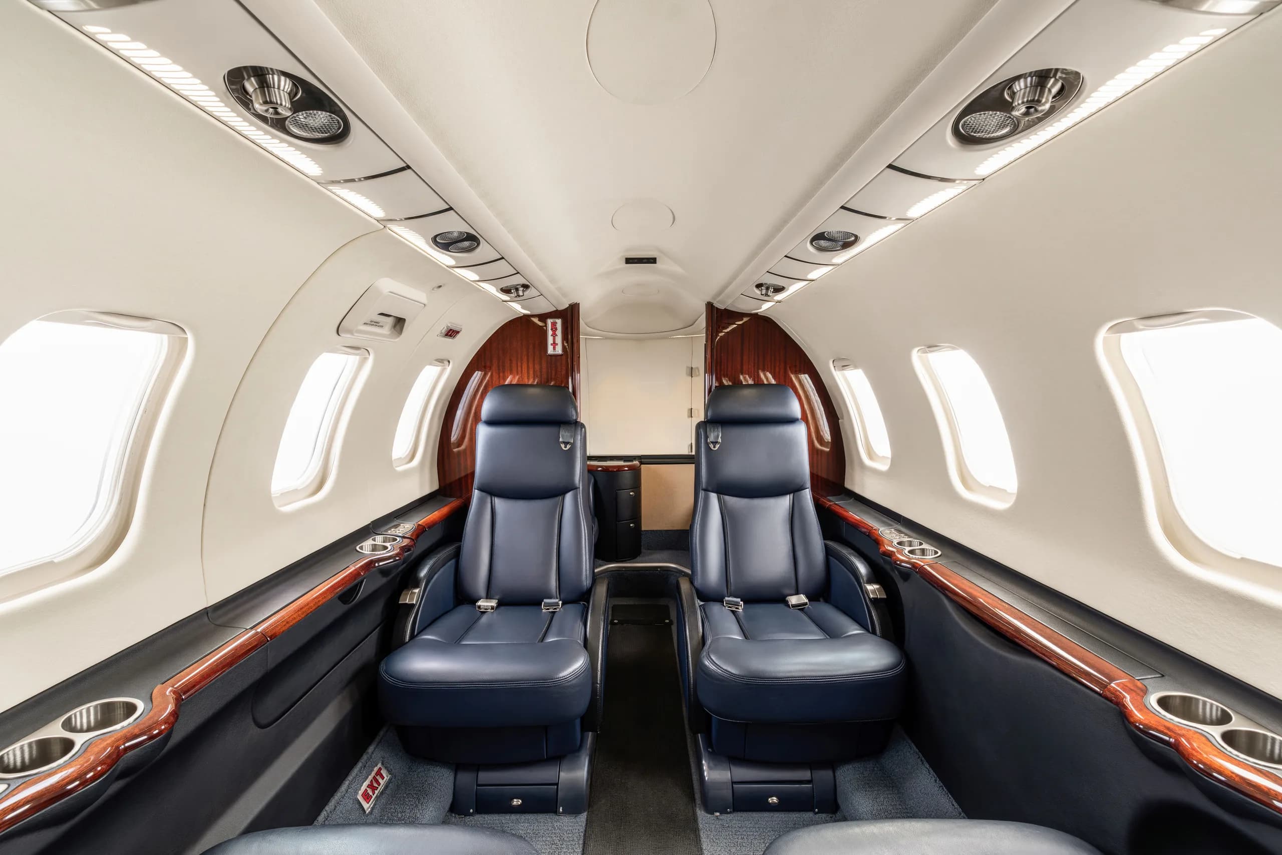 The interior cabin of a private jet, showing two rows of dark blue leather seats and light-colored walls with wood trim.