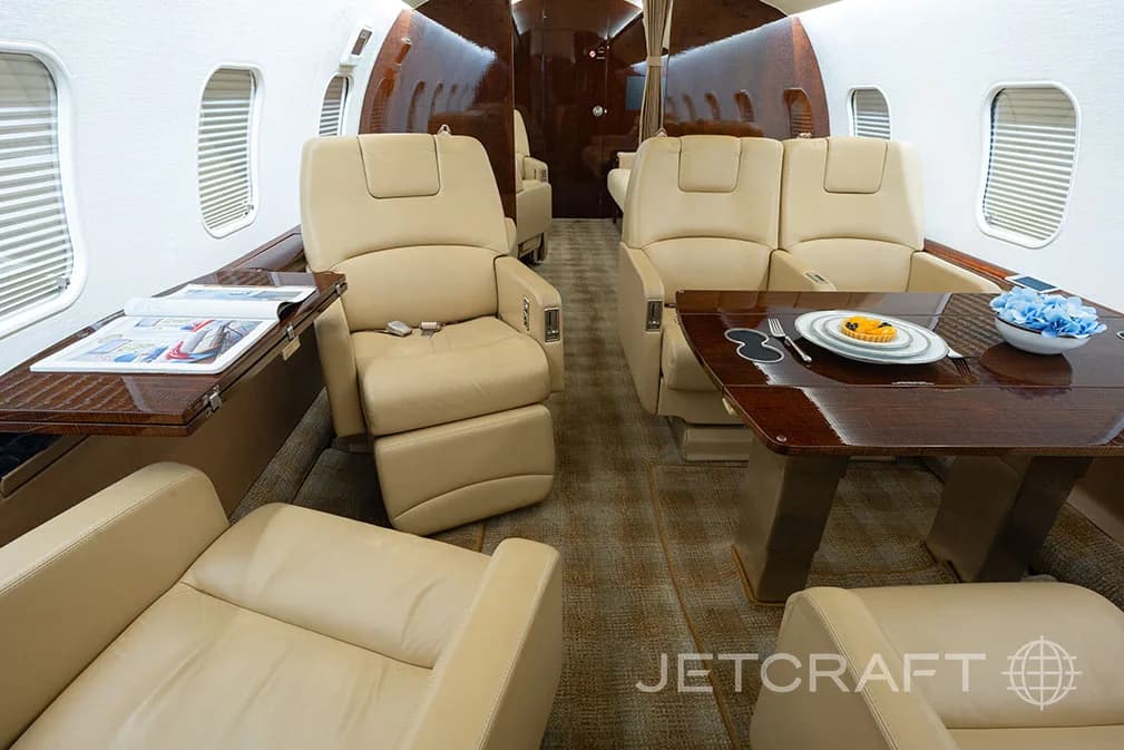 A private jet cabin interior with beige leather seats, a dining table set with a plate, and a side table with magazines.