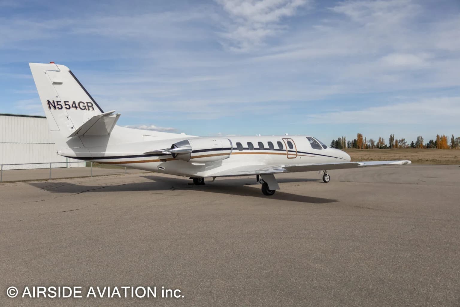 A white Cessna Citation business jet with gold and black stripes, registration N554GR, viewed from the rear-side on an airfield.