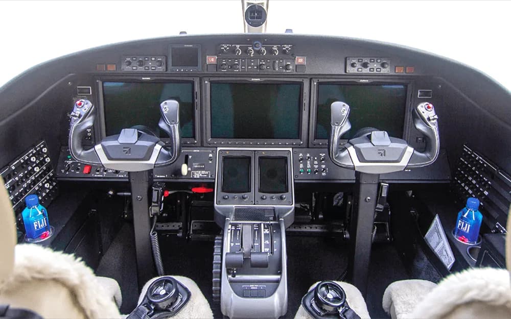 The cockpit of a jet features multiple large display screens, two control yokes, and a central throttle quadrant.
