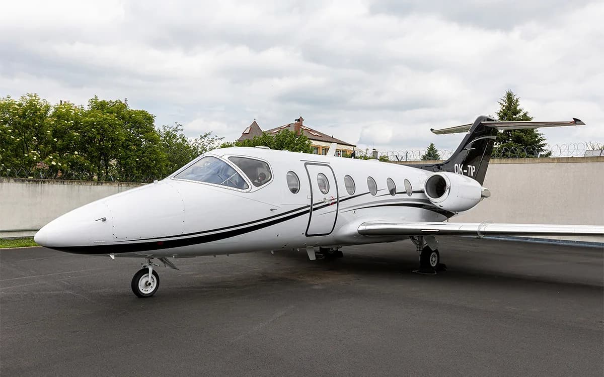 A white business jet with black and silver stripes and a black tail, registration OK-TIP, parked on an tarmac under a cloudy sky.