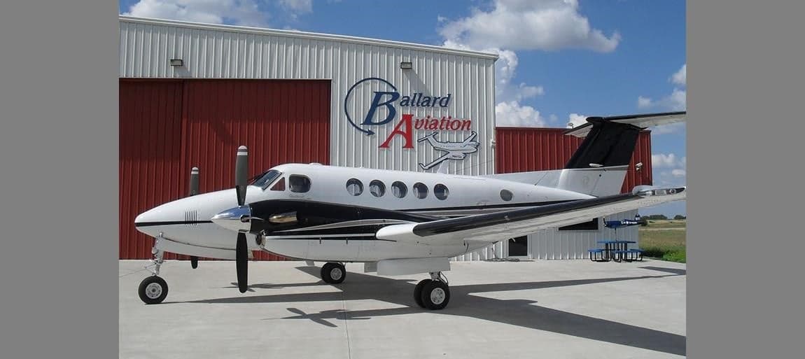 Side view of a King Air aircraft parked in front of a hangar with the Ballard Aviation logo.