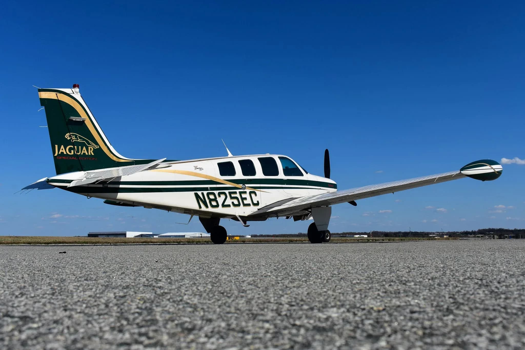 A low-angle side view of the N825EC Beechcraft Bonanza A36, featuring a white and green paint scheme with gold accents and a "Jaguar Special Edition" tail logo, parked on an airfield.