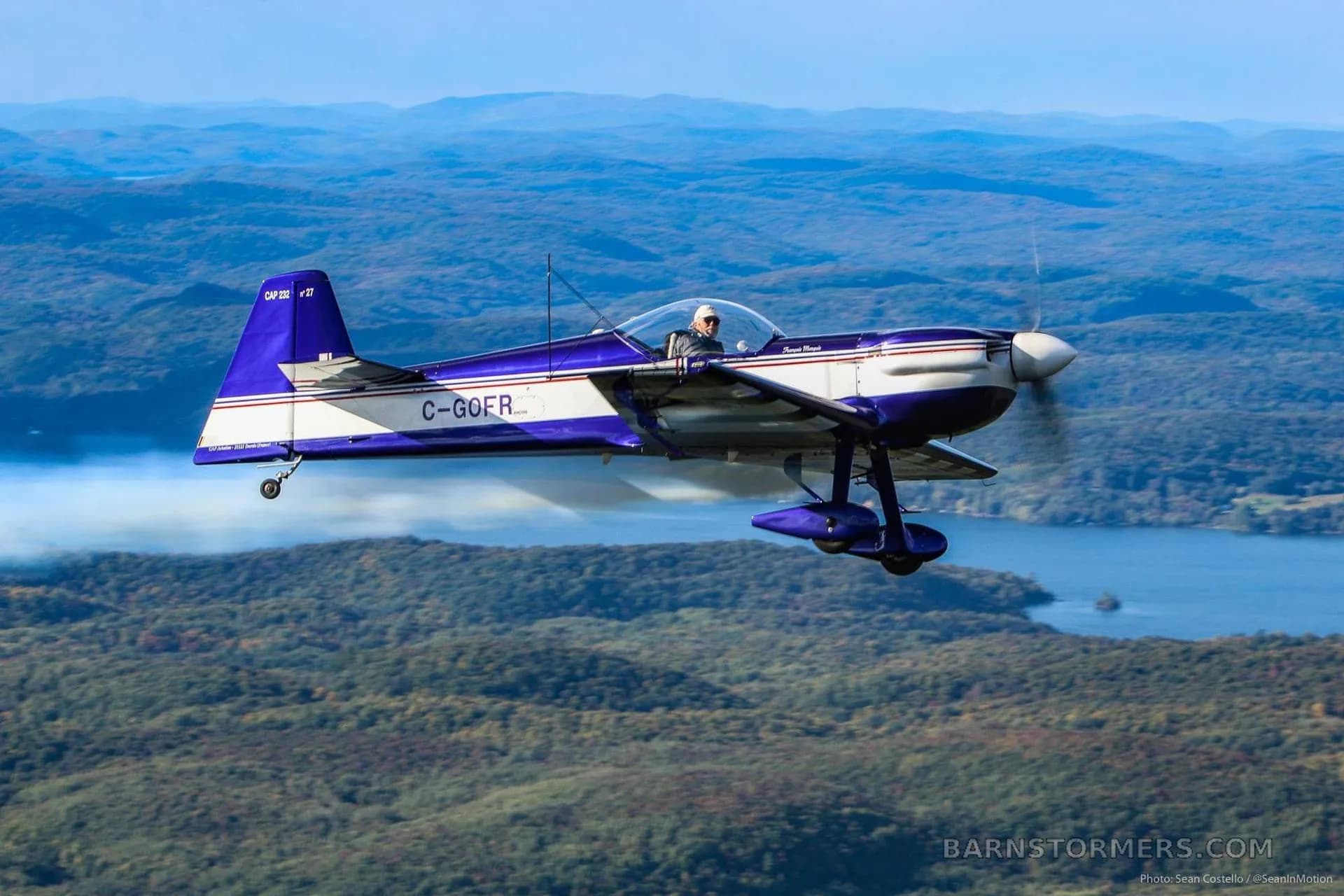 A blue and white CAP 10B aerobatic aircraft with registration C-GOFR flies over a mountainous, forested landscape with a lake, trailing white smoke.