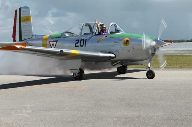 A silver single-engine propeller aircraft with green and yellow trim, with a pilot waving from the cockpit, on a runway.