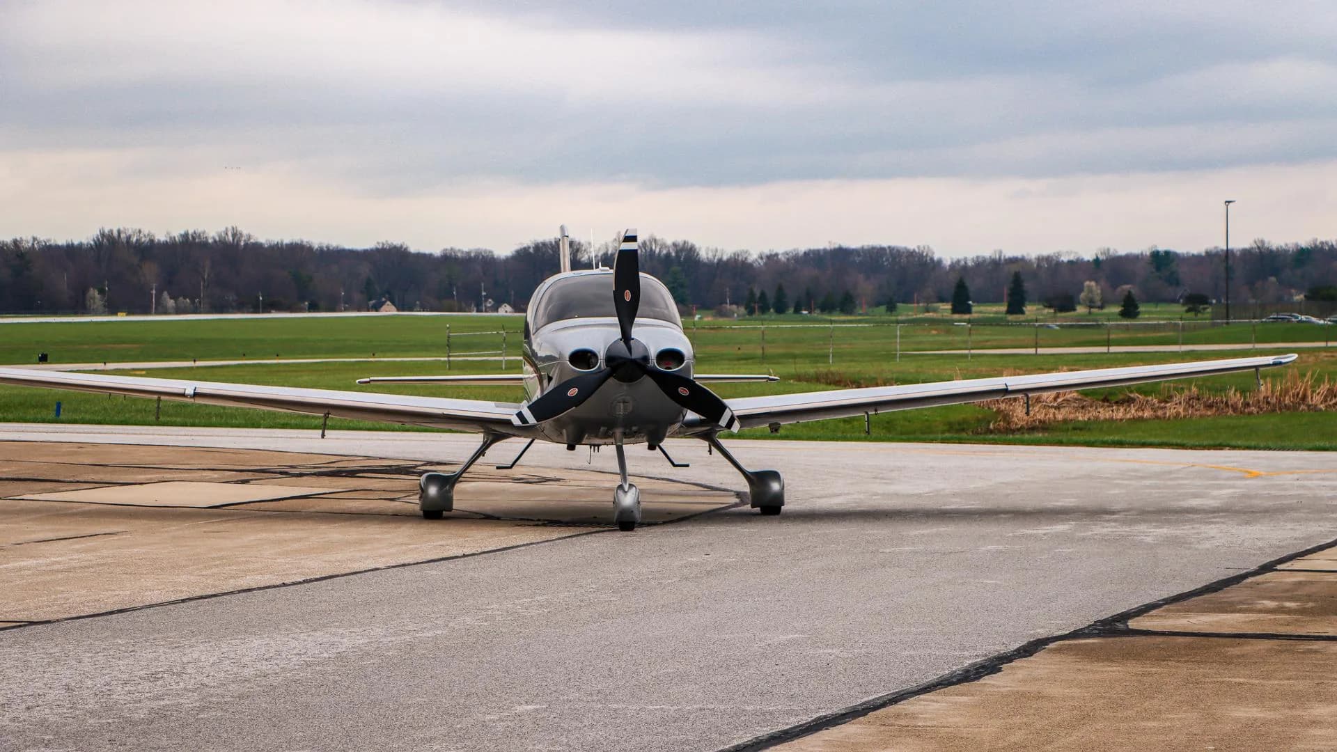 A silver and white Cirrus SR22 aircraft is seen from the front, parked on an airport tarmac under a cloudy sky.
