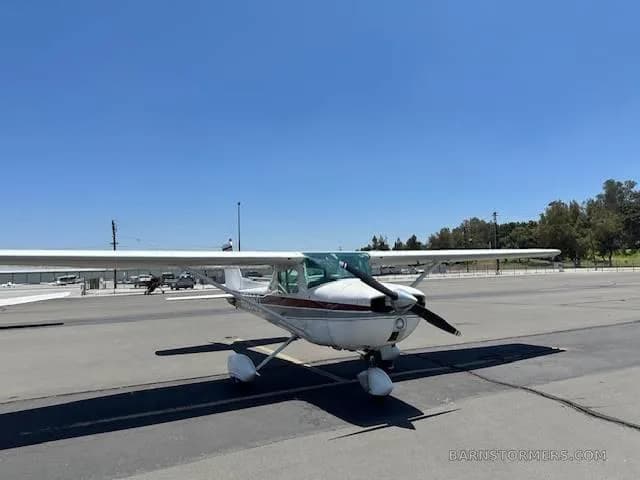 A white Cessna 172 with red and grey stripes is parked on an asphalt ramp, viewed from the front.