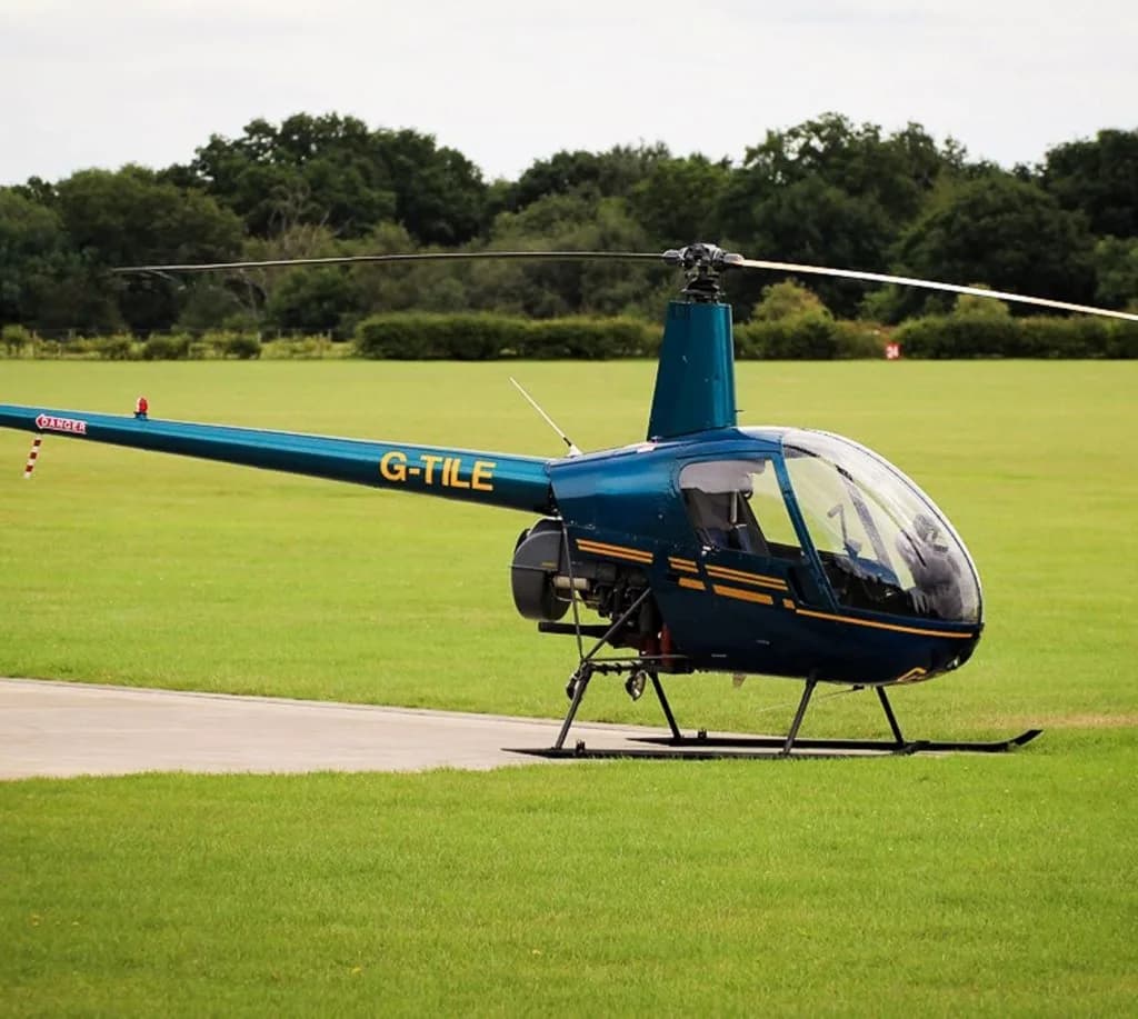 A teal Robinson R22 helicopter with yellow stripes parked on a concrete pad in a green field.