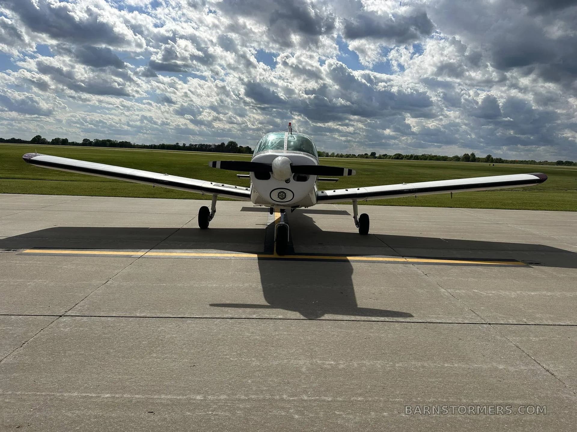 A white and maroon single-engine propeller aircraft is parked on an asphalt ramp, viewed head-on, under a partly cloudy sky.