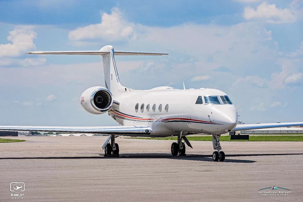 A front-quarter view of a white Gulfstream G550 aircraft with red and blue stripes, parked on an airfield.