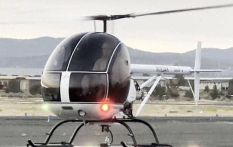 A white and blue helicopter sits on the runway with its lights on.