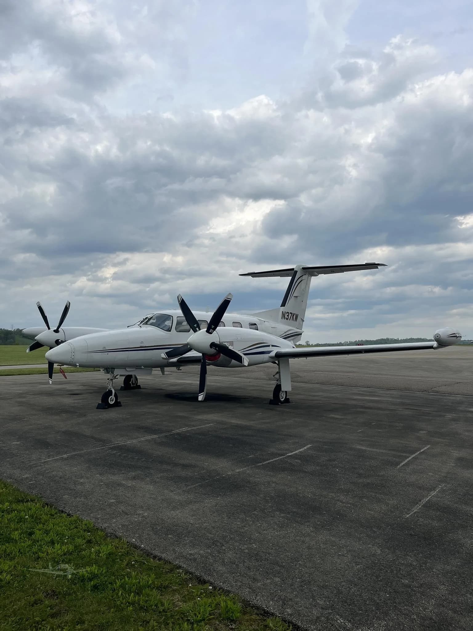 Three-quarter front view of a white twin-engine turboprop aircraft (N37KW) with black propellers, parked on an airfield under a cloudy sky.