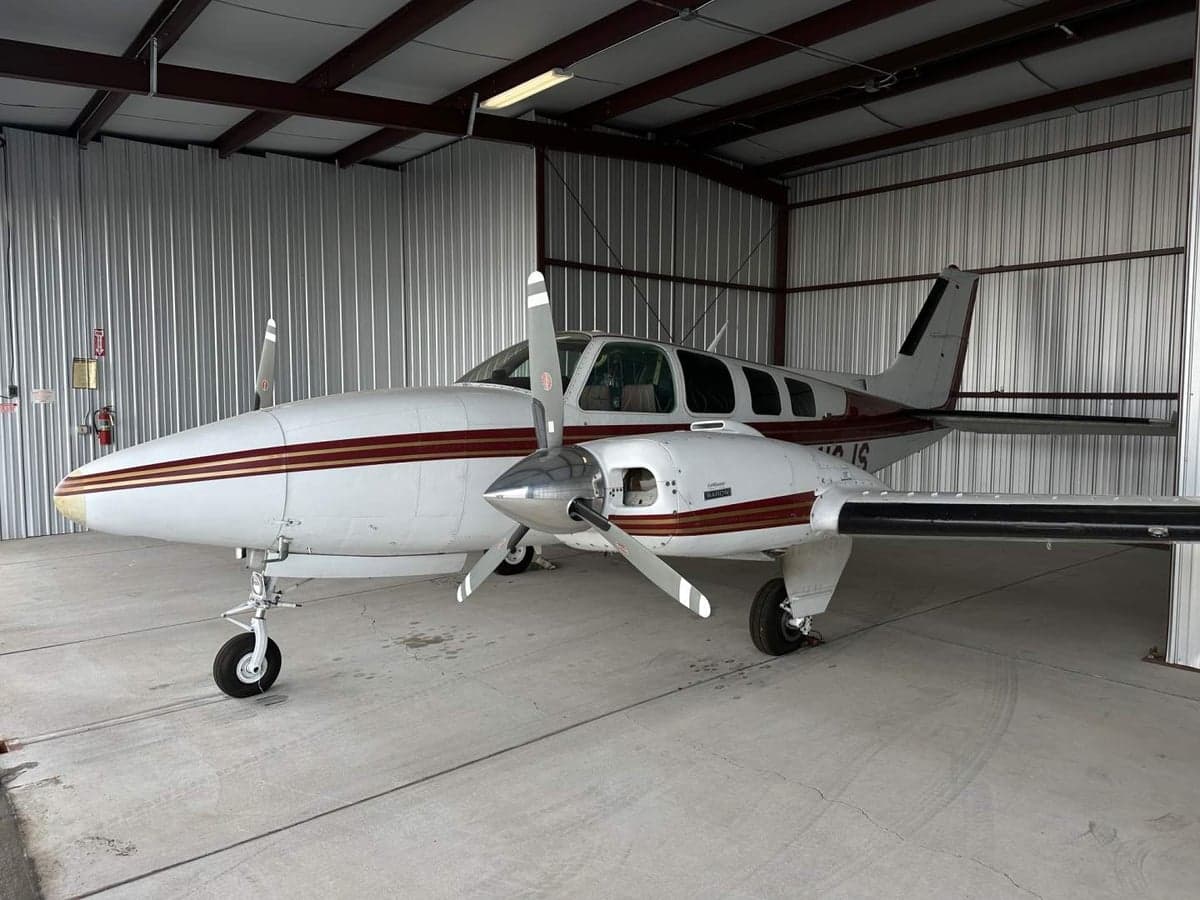 A Beechcraft Baron aircraft with a white and maroon paint scheme is parked inside a hangar.