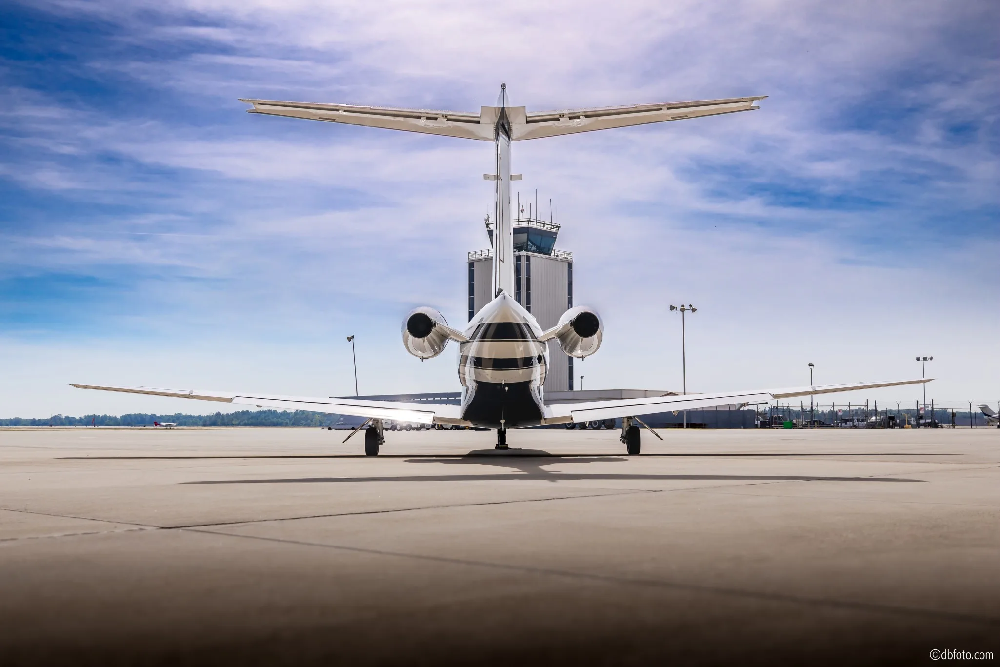 A rear view of a white Cessna Citation CJ3 business jet with blue and grey stripes, registration N5GU, parked on an airport tarmac with a control tower in the background.