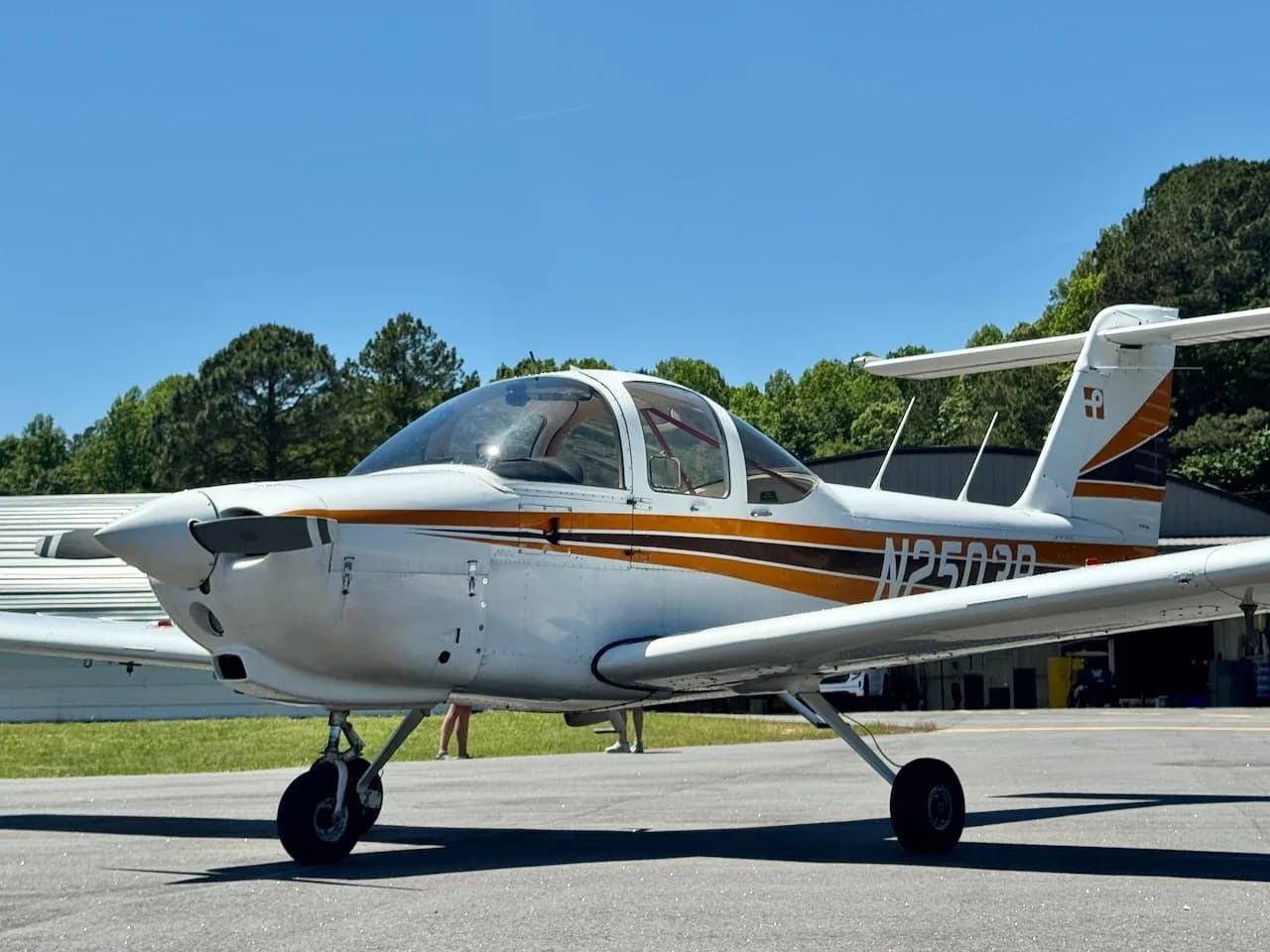 A white Piper PA-38 Tomahawk aircraft with orange and black stripes, seen from the front-left on an airfield.