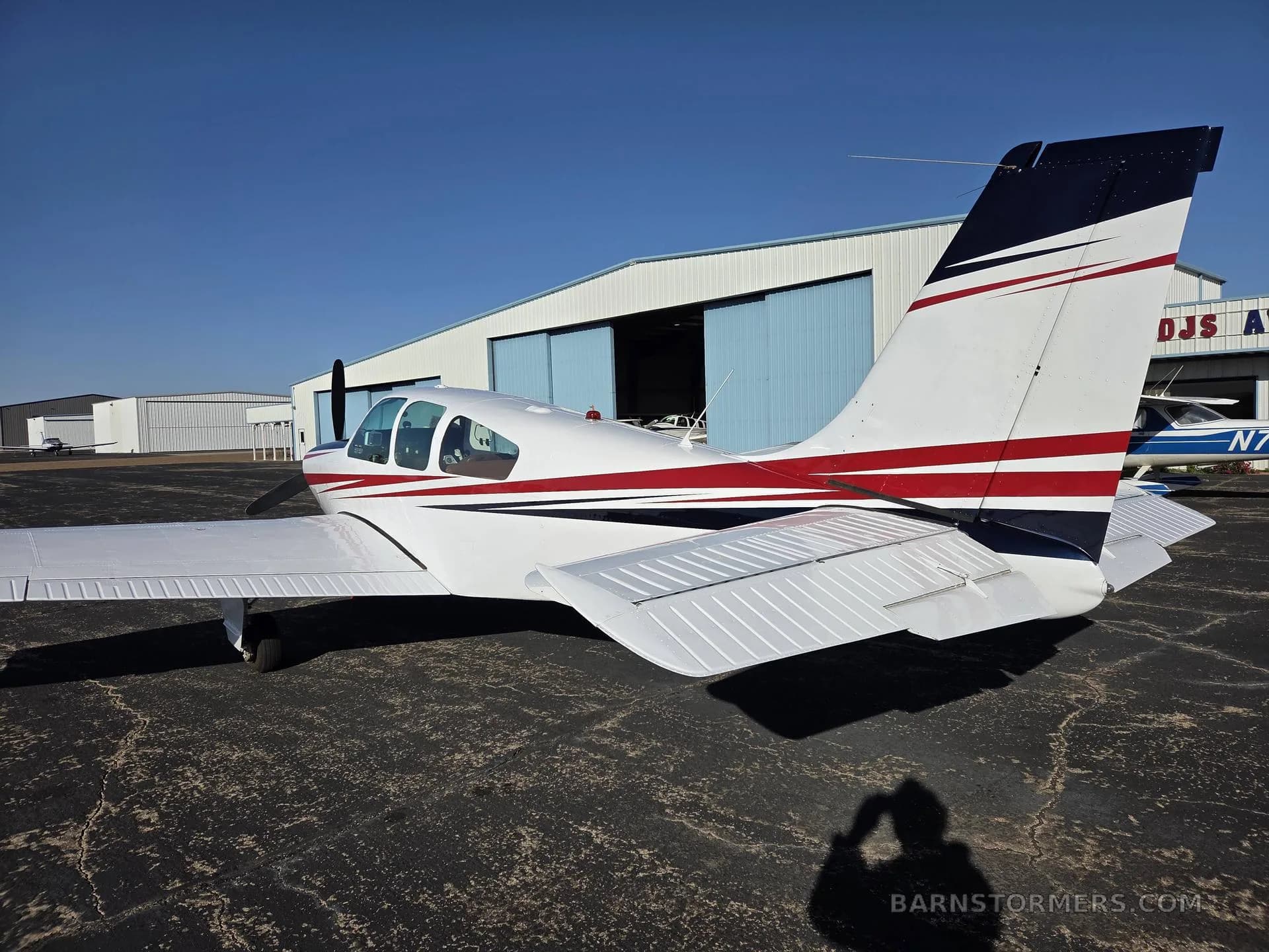 A rear-side view of a white single-engine aircraft with red and black stripes on the tail is parked on an asphalt tarmac in front of a hangar.