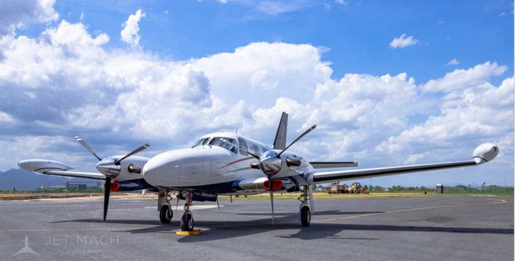 A white and blue twin-engine propeller aircraft with red accents sits on an airport tarmac under a blue sky.