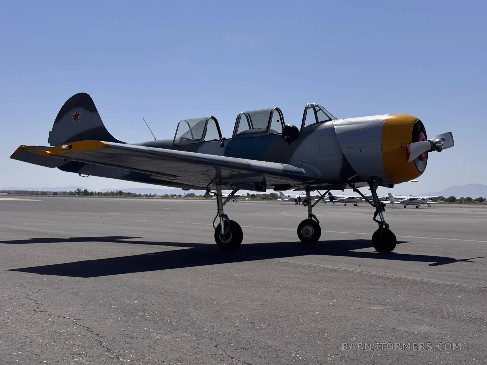 A side view of a blue and grey camouflage Yak-502GC aircraft with a yellow nose, parked on an airport tarmac under a clear blue sky.