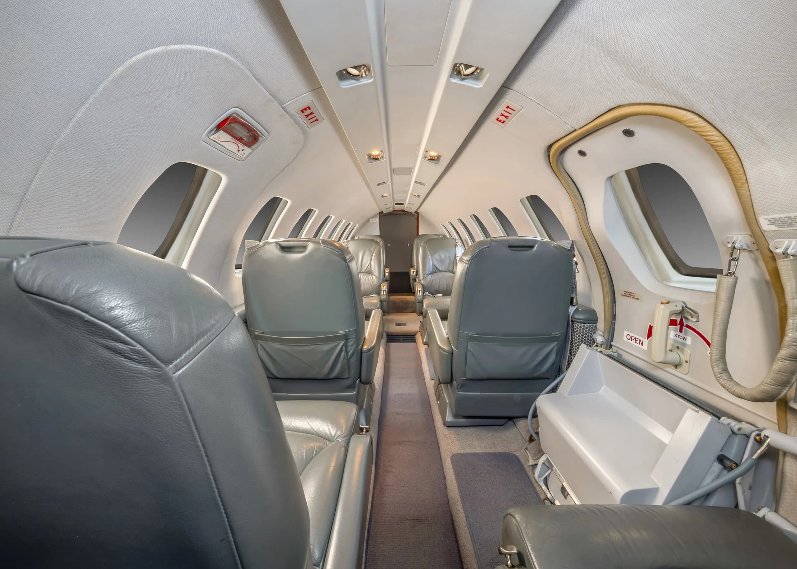 The aircraft cabin interior looking aft, featuring grey leather seats and an open entry door on the right.