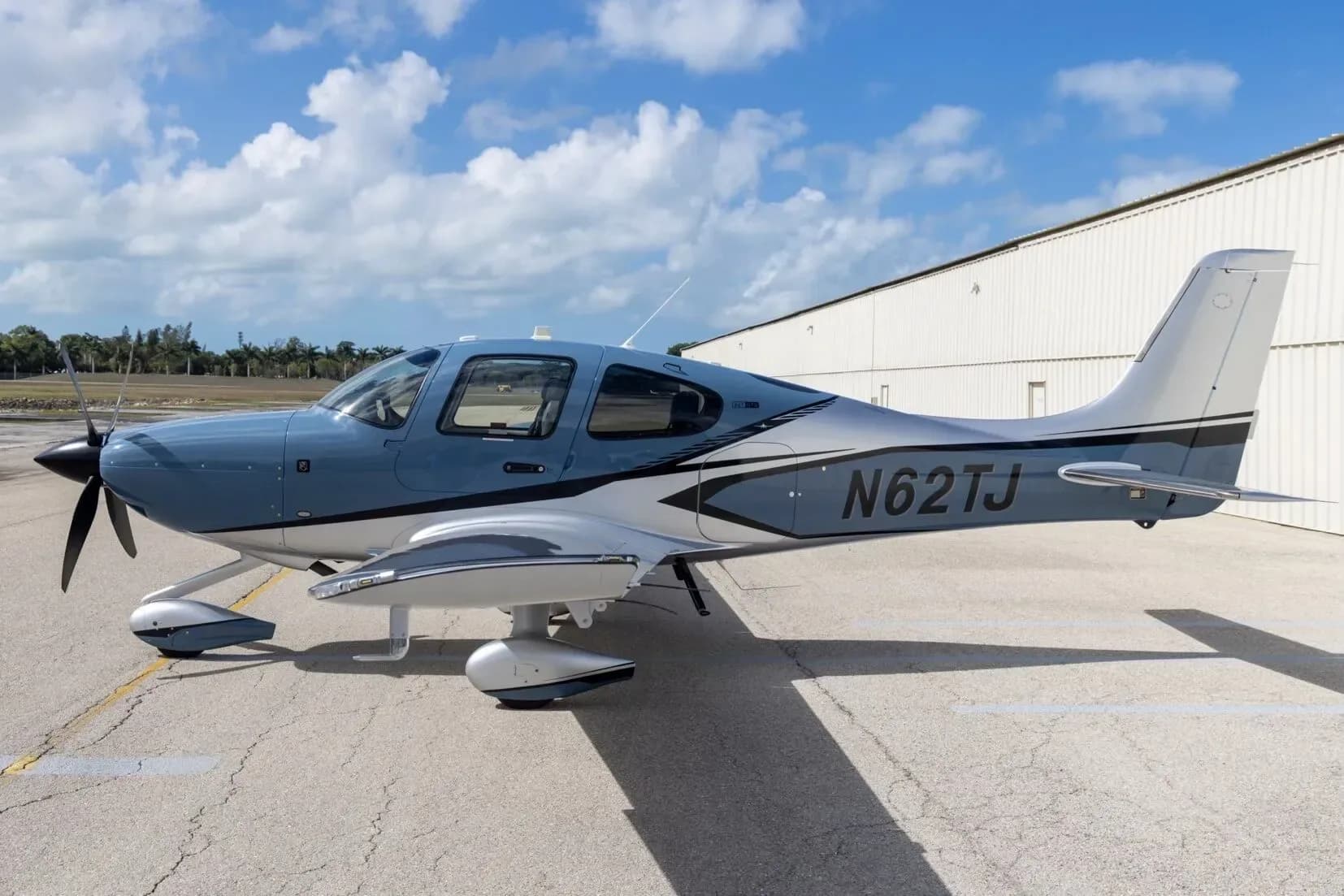 A blue and silver Cirrus SR22T aircraft with registration N62TJ parked on an airfield next to a white hangar.