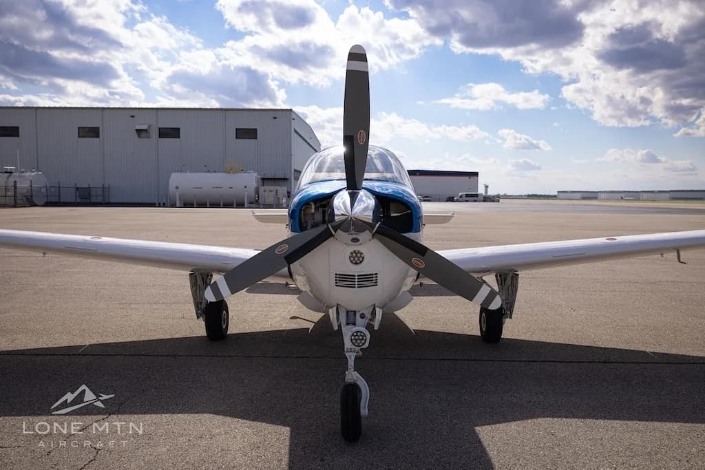 A head-on view of a white, blue, and red Beechcraft Bonanza N680JP with its propeller in front of a hangar.