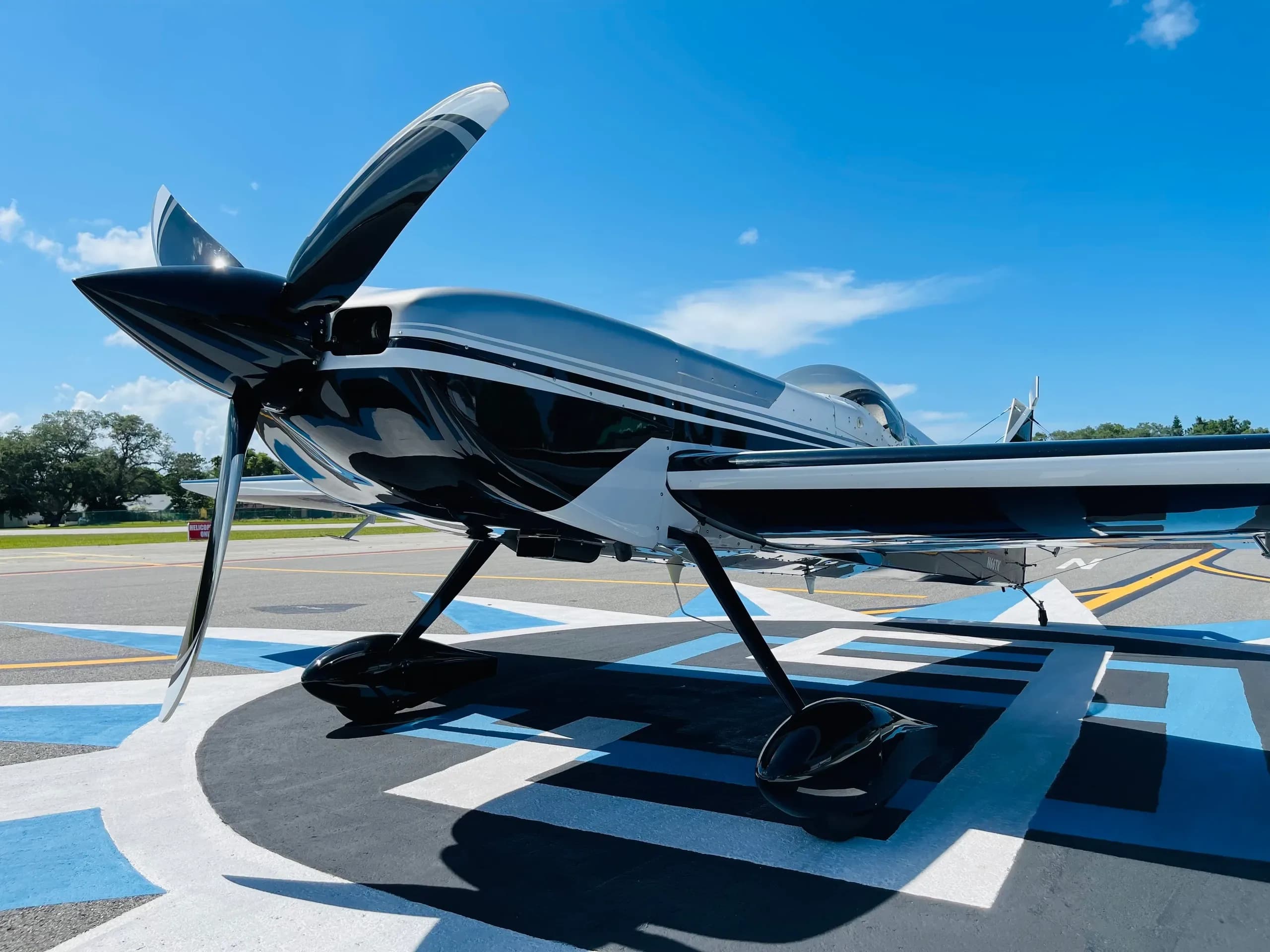 A close-up front view of a black and white aerobatic aircraft's propeller and nose on a sunny tarmac.