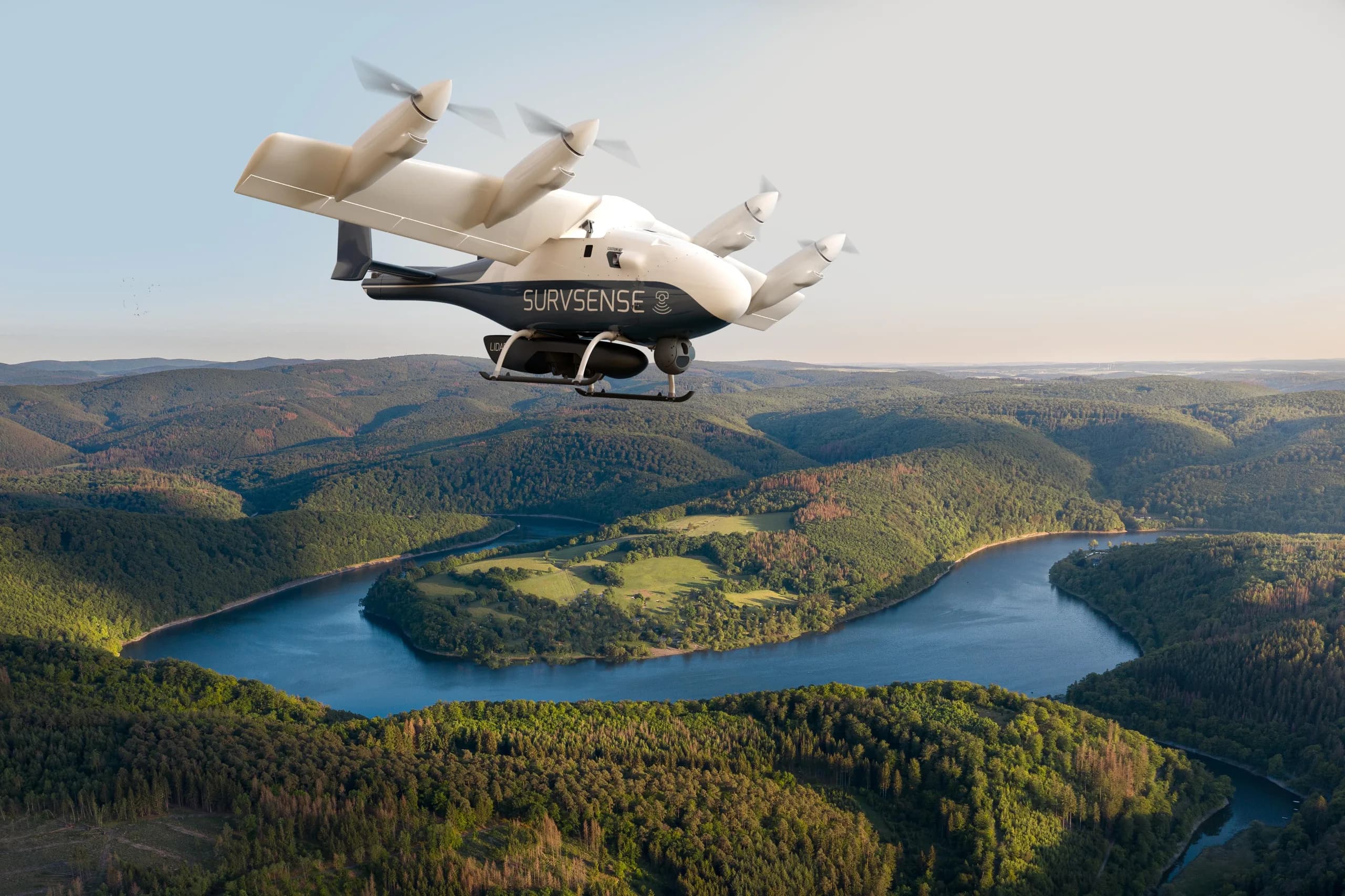 A dark blue and white SurvSense eVTOL aircraft flying over a green forested valley with a winding river.