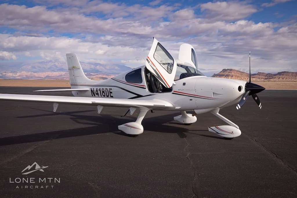 A white Cirrus SR22 aircraft with black and red stripes, registration N418DE, parked on an airfield with doors open.