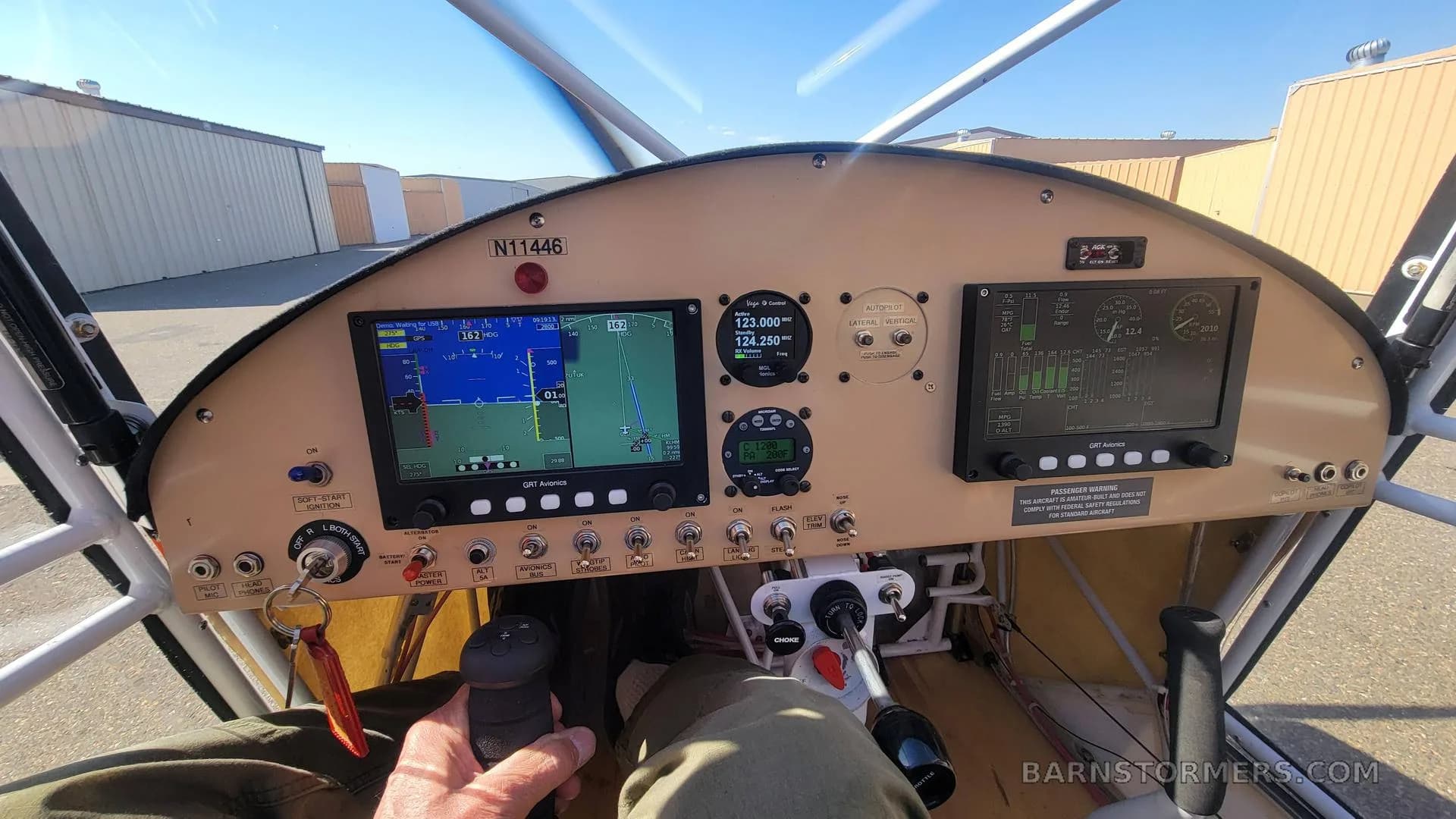 The cockpit instrument panel of the aircraft, featuring multiple digital displays, gauges, and controls, with a hand on the joystick.