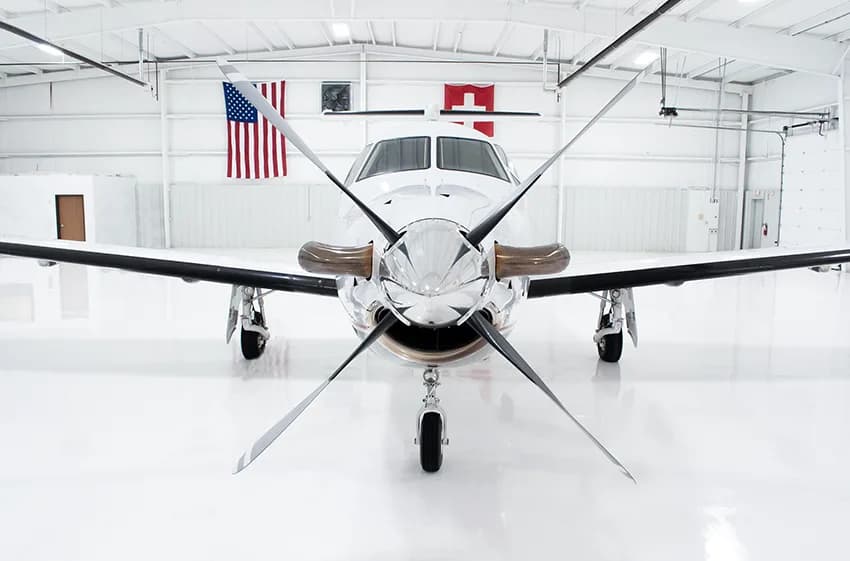 A side view of the fuselage of a white Pilatus PC-12NG aircraft, showing its gray and red stripe livery and cabin windows.