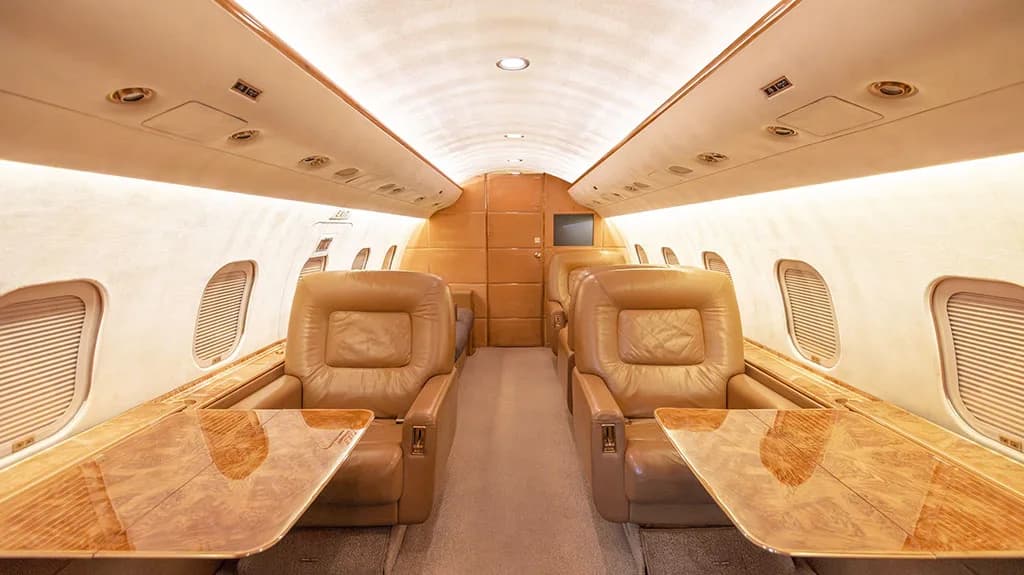 The private jet cabin interior showing two rows of tan leather seats with polished wooden tables extended.